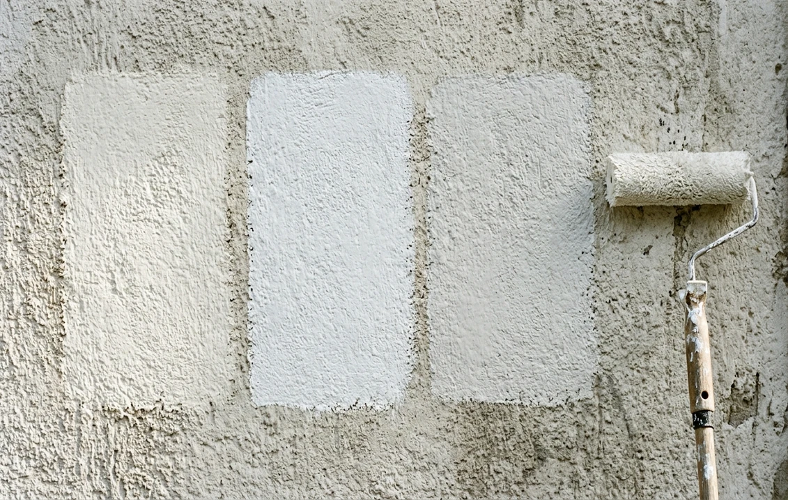 Drei Fassadenfarb-Musterflächen in leicht unterschiedlichen Weißtönen auf einer Putzwand neben einer Malerrolle