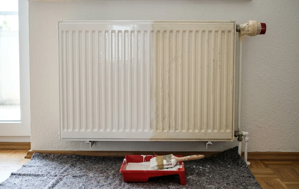 Weißer Flachheizkörper an der Wand, die linke Hälfte frisch glänzend weiß lackiert, die rechte Hälfte noch gelblich und stumpf, Thermostat abgeklebt, darunter Malervlies mit Farbwanne und Pinsel